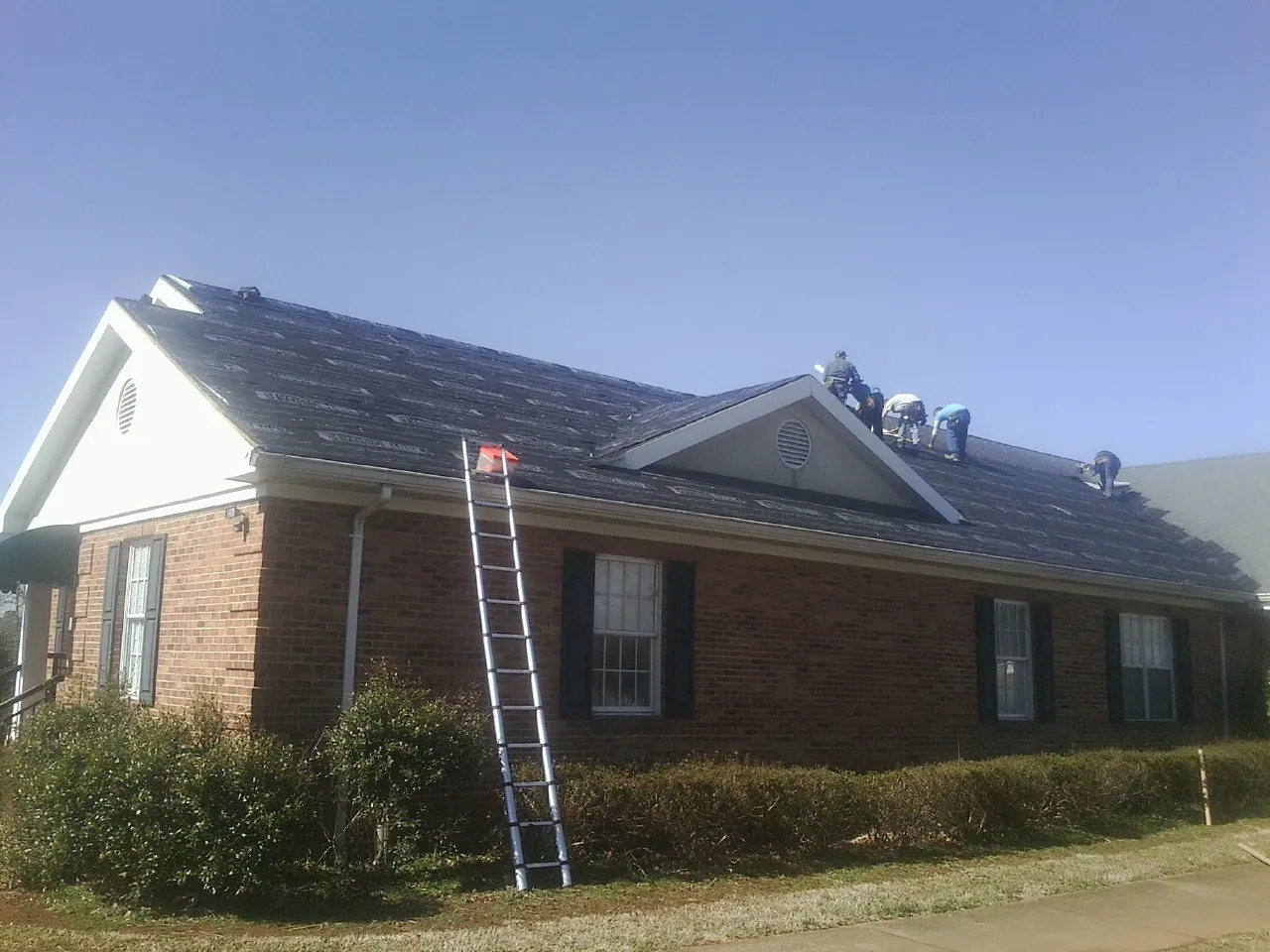 Crew tearing shingles off a brick ranch roof with a ladder set — Emergency Roof Repair work by our crew in Luxemburg