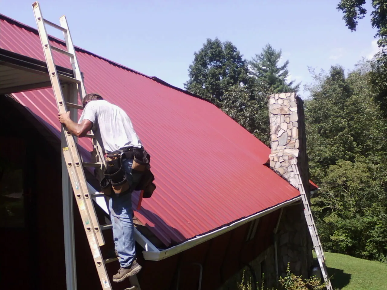 Roofer on a ladder beside a red metal roof and stone chimney — Soffit & Fascia Repair by Roofing Brothers in Luxemburg